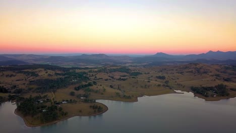 Rising-aerial-view-of-brown-hills-and-mountains-surrounding-lake-Moogera-at-sunset---Queensland,-Australia