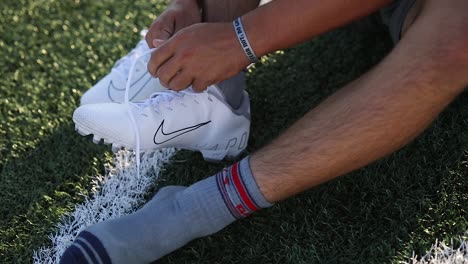 Slow-Motion-Shot-of-an-American-Football-Player-sitting-on-the-football-field-and-putting-on-and-tying-his-Nike-Cleats
