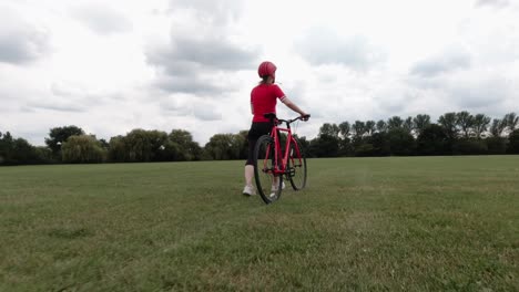 Caucasian-female-cyclist-in-red-top-walking-with-her-bike-in-a-park