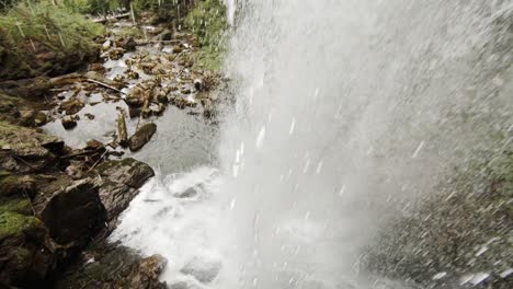 Camera-showing-and-moving-behind-a-waterfall,-in-daylight,-in-mountain-region