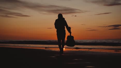 Man-running-with-guitar-in-back-sand-beach-at-sunset