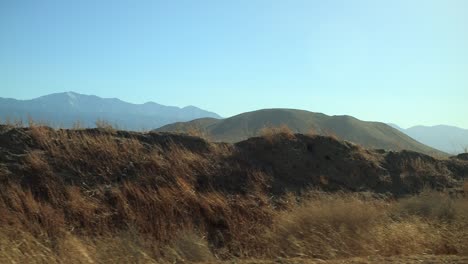 Driving-through-the-empty-desert-with-dry-plants-passing-by-on-the-side-of-the-road-with-hills-and-mountains-in-the-distance,-closer