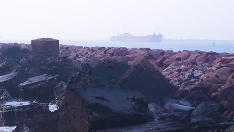 Large-cargo-ship-entering-the-Port-of-Liepaja-in-foggy-day,-stone-pier-in-foreground,-waves-splashing,-distant-medium-shot