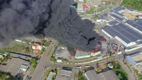 Dense-and-toxic-black-smoke-rising-from-burning-warehouse,-scenic-shot-with-drone