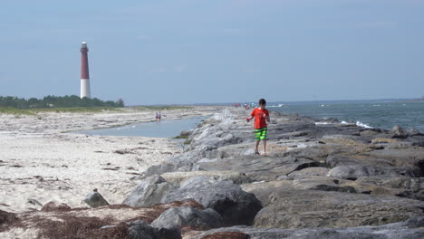 Barnegat,-New-Jersey---August-14,-2020:-A-boy-walking-on-the-rock-jetty-at-Barnegat-jetty-with-the-lighthouse-in-the-background