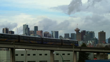 Filmische-Weite-Aufnahme-Des-Vorbeifahrenden-öffentlichen-Zugs-Auf-Der-Brücke-Und-Der-Skyline-Der-Metropole-Vancouver-Im-Hintergrund-Während-Des-Bewölkten-Tages