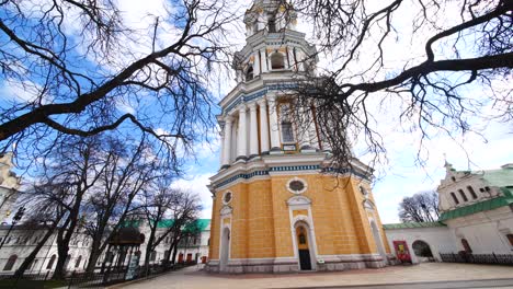 Yellow,-blue-and-white-baroque-bell-tower-behind-brown-winter-trees-without-leaves,-panning-down,-Kyiv-Ukraine,-Kiev-Pechersk-Lavra