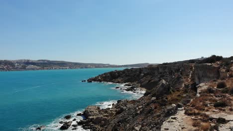Aerial-forward-horizon-view-of-rocky-cliffs,-coast-with-calm-blue-sea-in-Mellieha-bay,-Malta
