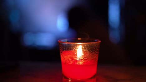 Red-candle-at-night-burning,-flickering-in-glass-jar-on-marble-table-with-large-tv-television-on-and-people-moving-around-in-background-silhouette---in-4K-30fps-