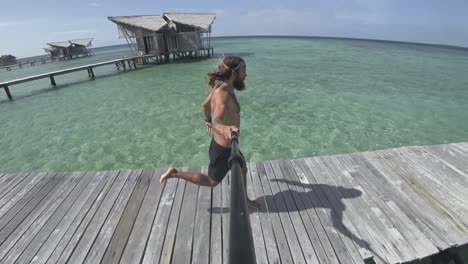 A-young,-fit-and-strong-man-is-running-on-the-wooden-jetty-in-the-sun-while-filming-himself-with-a-selfie-stick-and-then-runs-into-his-luxury-bungalow-sitting-on-top-of-the-water