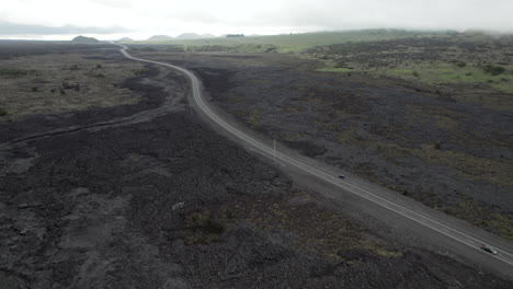 Aerial-tilt-up-shot-of-driving-vehicles-on-abandoned-road-surrounded-by-dried-volcanic-lava-landscape-and-cloudy-day