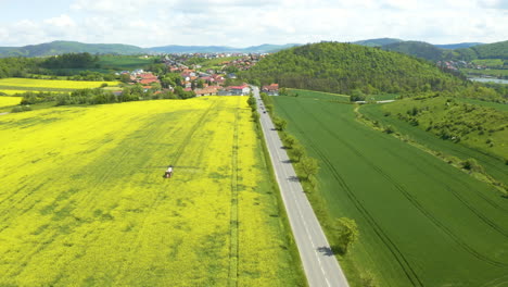 Tractor-spraying-pesticide-in-a-rapeseed-field-by-a-road,car-passing