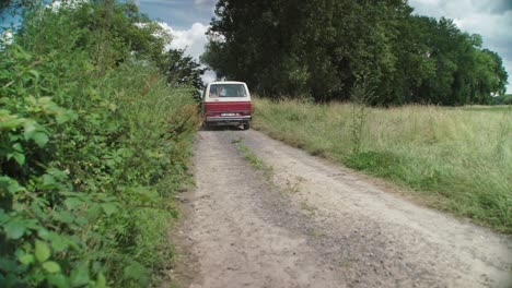 White-red-retro-T3-van-driving-away-on-rural-dirt-road,-behind-shot