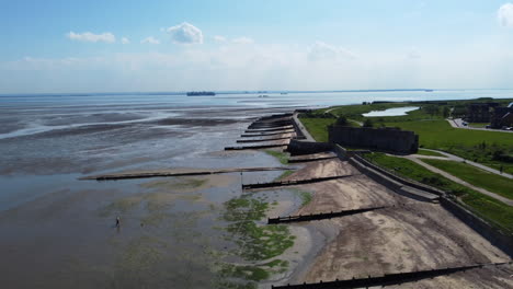 Drone-flight-over-estuary-mudflats-and-a-beach-separated-by-old-wooden-groynes-at-low-tide,-with-a-seawall-and-an-abandoned-military-garrison-on-the-right-hand-side,-on-a-clear-and-hot-summer-day