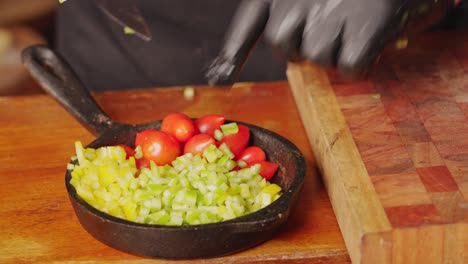 Preparation-of-salad-ingredients,-professional-chef-wearing-gloves,-transferring-diced-green-bell-pepper-into-stone-pan-next-to-juicy-cherry-tomato
