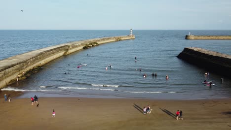 People-having-fun-on-the-beach-and-swimming-in-the-sea,-aerial-pan-view-of-harbour-entrance-on-a-bright-sunny-day-during-summertime