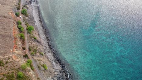 Aerial-drone-of-white-car-moving-slowly-along-a-rough-bumpy-coastal-road-with-turquoise-ocean-in-Timor-Leste