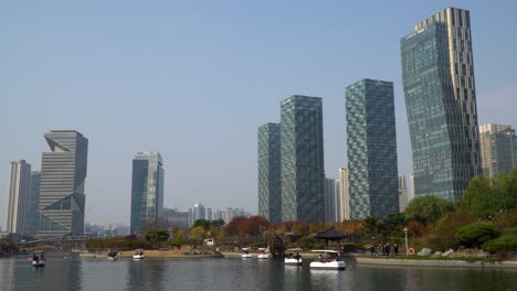 Städtische-Skyline-Von-Incheon-Songdo-Central-Park-Mit-Menschen,-Die-In-Kleinen-Familienbooten-Auf-Einem-See-Boot-Fahren