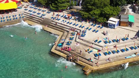 Aerial-View-Of-People-Swims-At-Adriatic-Sea-With-Lounge-Chairs-At-Novi-Beach-In-Novi-Vinodolski,-Croatia