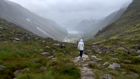 Rear-view-of-female-person-with-raincoat-hiking-on-rocky-path-between-giant-mountains-of-Ireland-during-foggy-day-in-nature---Slow-motion-wide-shot