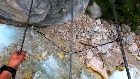 POV-shot-of-a-men-balancing-across-a-crystal-clear-river-on-a-steel-cable-bridge
