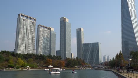 Songdo-Central-Park-in-Incheon,-South-Korea-with-people-enjoying-traveling-by-boats-and-walking-by-the-lake-with-high-rise-skyscrapers-against-clear-sky,-Posco-Tower---establishing-shot