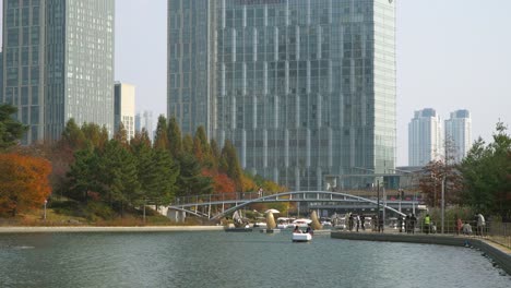 People-doing-water-activities-and-walking-by-the-lake-in-Songdo-Central-Park-in-Incheon-with-modern-Highrise-buildings-on-background---establishing-shot,-South-Korea