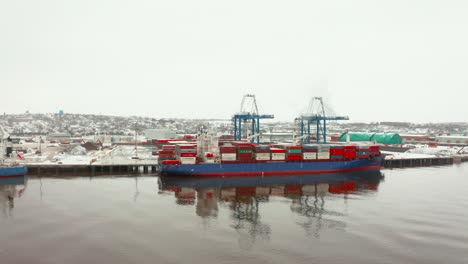 Low-aerial-view-of-a-cargo-ship-docked-in-port-on-a-snowy-winter-day