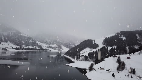 Foggy-mountain-and-lake-water-reflection-in-winter-landscape-during-snowfall,-aerial-drone-view