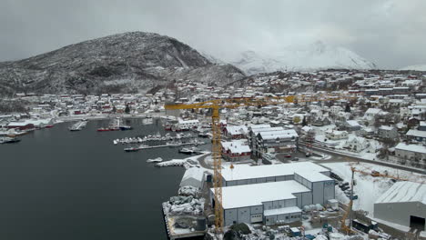 Drone-orbit-around-yellow-crane-at-harbour-of-fishing-town-covered-in-snow,-Arctic