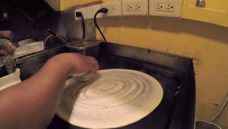 Indian-dosa-pancake-mix-being-spread-and-cooked-on-hot-plate-in-kitchen
