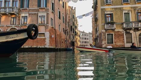 Water-surface-pov-of-boat-passing-with-tourists-on-board-and-rowing-man-with-hat-standing,-hanging-sheets-in-background,-Venice-in-Italy
