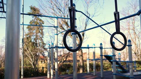 gymnastic-rings-move-in-the-wind-with-trees-and-calisthenics-structure-on-the-background,-steady-close-up-shot