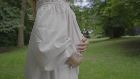 Closeup-shot-of-a-pregnant-young-woman-wearing-a-beautiful-white-dress-and-holding-her-large-belly,-while-walking-through-her-lush-green-garden