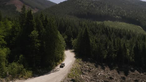 Aerial-pan-left-of-a-jeep-driving-on-a-dusty-hillside-road-surrounded-by-dense-green-pine-forest,-mountains-in-background,-British-Columbia,-Canada