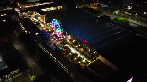 Night,-aerial-and-circular-view-of-a-shopping-center-with-an-amusement-park-with-colored-lights-on-the-terrace-and-people-enjoying