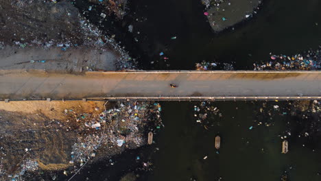 Aerial-top-down-of-old-narrowed-suspension-bridge-with-scooter-driving-over-a-polluted-river-filled-with-garbage-plastic-waste-pollution-trash