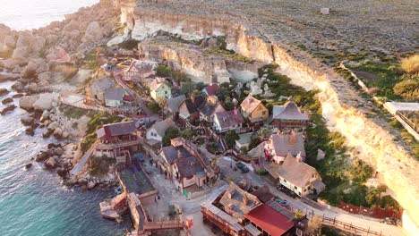 Rocky-cliff-side-coastal-wooden-town-in-Malta-island,-aerial-view-during-golden-sunset