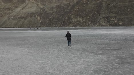 Macho-Adulto-Caminando-Por-El-Lago-Khalti-Congelado-Con-Gente-Patinando-Sobre-Hielo-En-Segundo-Plano