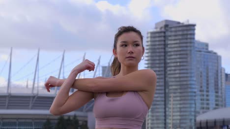 Woman-stretching-triceps-with-arm-across-body,-city-buildings-in-background