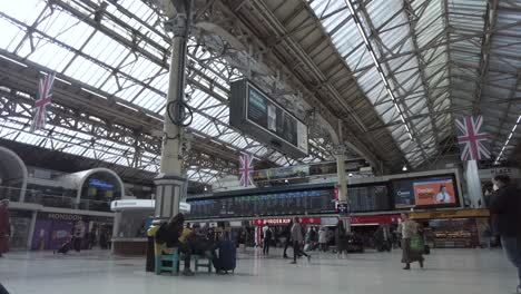Innenansicht-Der-Victoria-Station-In-London-Mit-Britischer-Flagge,-Die-Vom-Dach-Hängt,-Und-Pendlern,-Die-Vorbeigehen