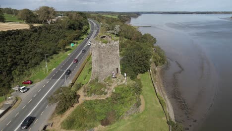 Drone-shot-of-a-busy-road-and-old-castle-ruins-with-a-wide-river-estuary-in-the-background
