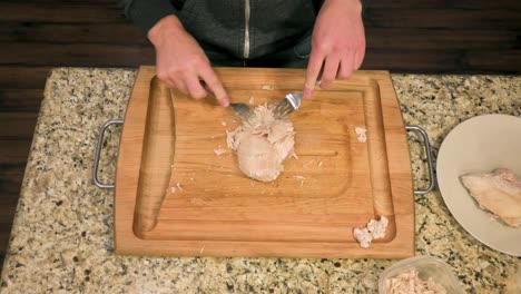 Caucasian-male-shredding-skinless-steamed-chicken-breast-with-forks-on-a-wooden-cutting-board