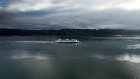 Slowly-tracking-a-commuter-ferry-as-it-cruises-along-a-calm-waterway-reflecting-dark-gloomy-clouds,-aerial