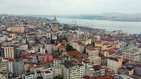 Aerial-drone-revealing-Galata-Tower-in-surrounded-by-old-European-residential-buildings-on-a-cloudy-day-in-Istanbul-Turkey-with-the-Bosphorus-River-in-the-distance