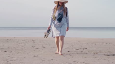 Caucasian-woman-walking-towards-camera-at-sea-wears-summery-clothes-and-hat