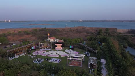 aerial-footage-of-an-empty-event-space-decorated-right-by-the-ocean,-pastel-colors-event-set-up,-blue-clear-sky,-blue-ocean,-visible-stage-area-and-chair-set-ups,-skyline-in-the-distance,-ocean-views