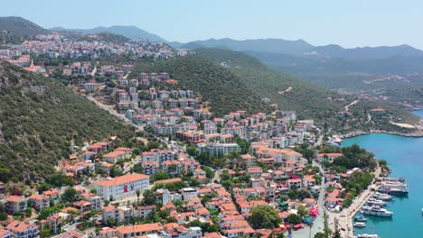 Wide-aerial-drone-of-European-buildings-with-orange-roofs-on-a-large-coastal-hill-and-boats-docked-at-the-marina-in-Kas-Turkey-on-a-sunny-summer-day