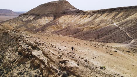 Aus-Der-Luft-Herauszoomen-Vom-Wanderer,-Der-Auf-Dem-Mount-Ramon-Desert-Trail-Winkt-Und-An-Einem-Sonnigen-Tag-Berggipfel-Und-Krater-Zeigt