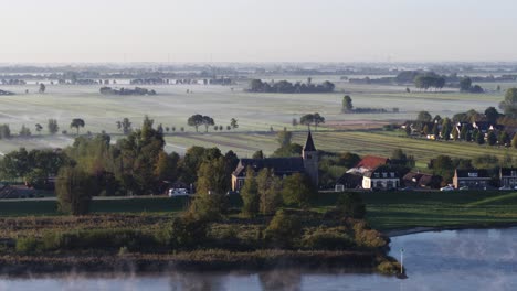 Small-riverside-town-with-church-tower-in-Netherlands-during-rising-fog,-aerial-view